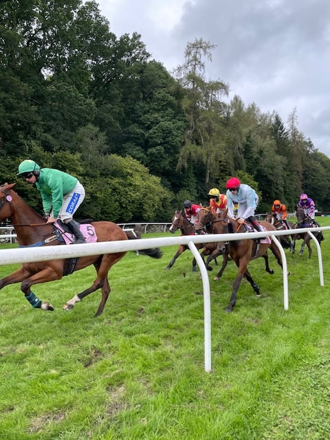 Horses running past with jockeys on their backs in colourful outfits. Cartmel racecourse track